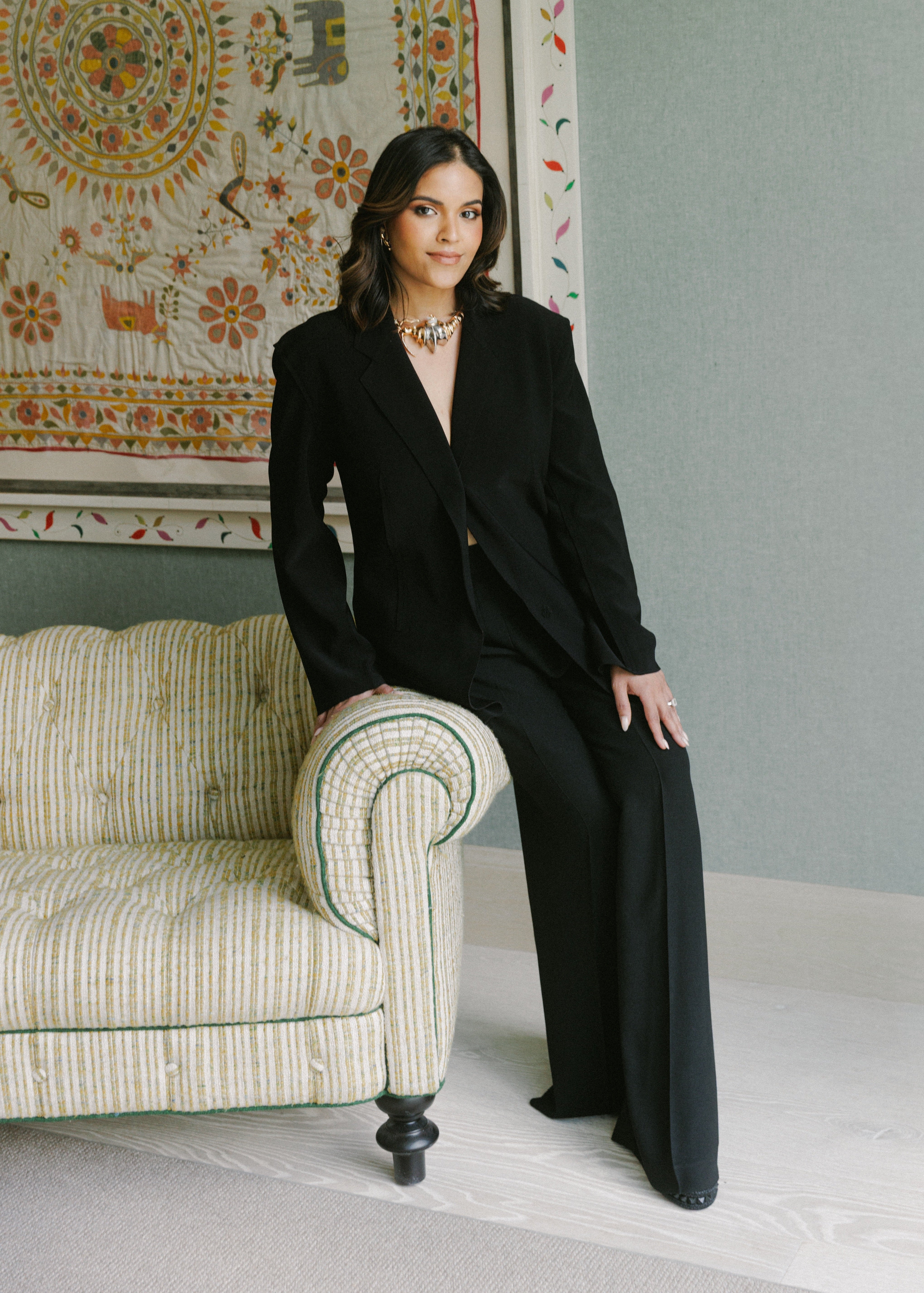 Archana Yenna, founder and CEO of Sincerely India, in a black tailored suit and sculptural gold necklace, seated before an embroidered wall textile